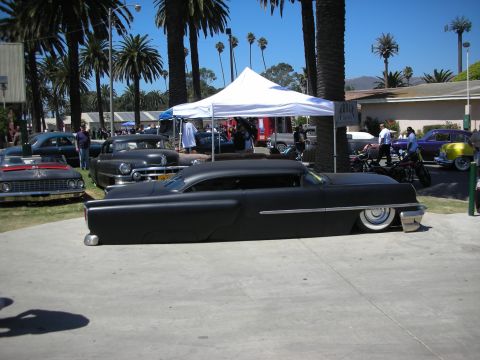 1998 Ford Crown Victoria - Side profile of a chopped, slammed flat-black 1950s custom with whitewall tires parked among other classics under palms at a show.