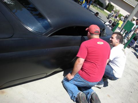 1998 Ford Crown Victoria - Two men crouch and inspect the front fender and headlight area of a flat-black chopped custom at an outdoor show.