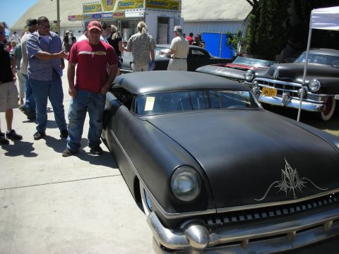 1998 Ford Crown Victoria - chopped flat-black mid-1950s custom coupe with delicate pinstriping and a man posing beside it among other classics at a show.