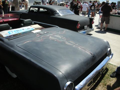 1998 Ford Crown Victoria - flat-black custom car's hood decorated with red-and-white flame pinstriping, parked beside other customs in a crowded show lot.