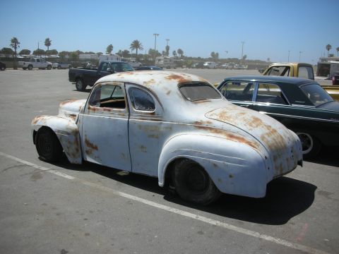 1998 Ford Crown Victoria - weathered vintage coupe in gray primer with surface rust sits in a paved lot with palm trees and other classic cars in the background.