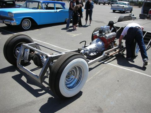 1998 Ford Crown Victoria - bare hot rod frame fitted with a chrome V8 engine and whitewall tires sits on a trailer while a man leans over it at a car show.