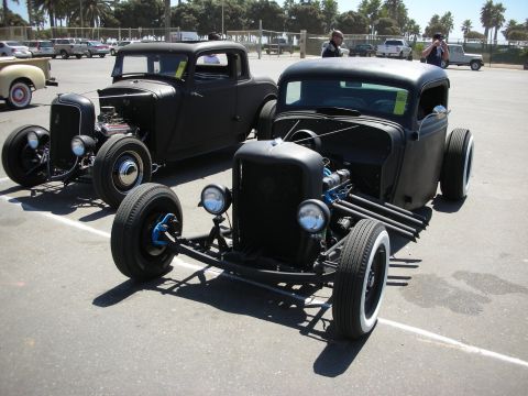 1998 Ford Crown Victoria - Two black rat-rod pickups with exposed frames, blue spoke wheels, and whitewall tires sit side by side in a paved lot near palm trees.