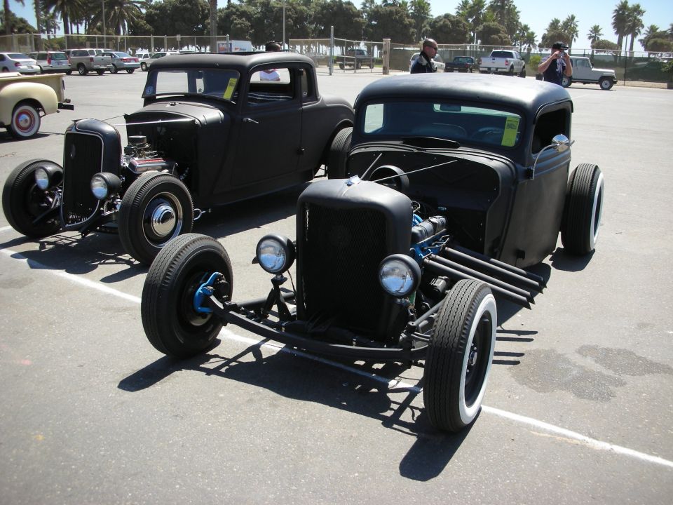 Two black channeled rat-rod trucks parked side by side in a beach lot