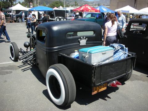 1998 Ford Crown Victoria - Rear view of a black rat-rod pickup with whitewall tires, a vintage cooler in the bed, and a California license plate at a busy outdoor sho…