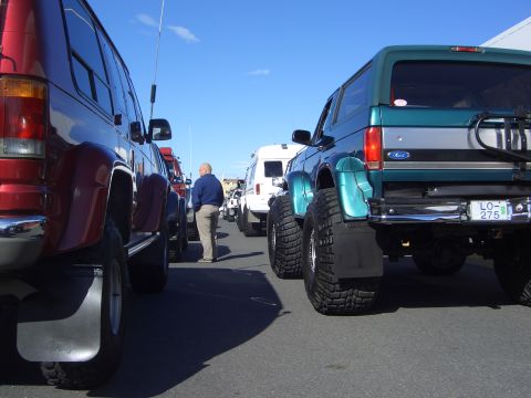 1996 Ford Bronco - A line of modified SUVs and vans are parked on a sunny day, with a teal Ford Bronco prominent in the foreground.