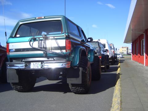 1996 Ford Bronco - A line of SUVs including a green Ford Bronco are parked next to a red building.