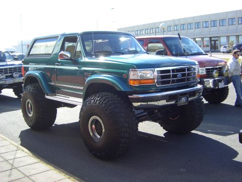 1996 Ford Bronco - This lifted 1996 Ford Bronco features large off-road tires and a distinctive green paint job, parked alongside other vehicles.