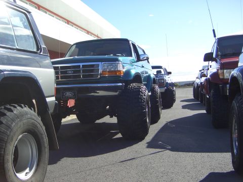 1996 Ford Bronco - Several modified trucks and SUVs parked in a line under a clear sky.