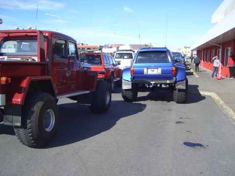 1996 Ford Bronco - Two modified trucks are parked in a lot next to a building with people walking by.