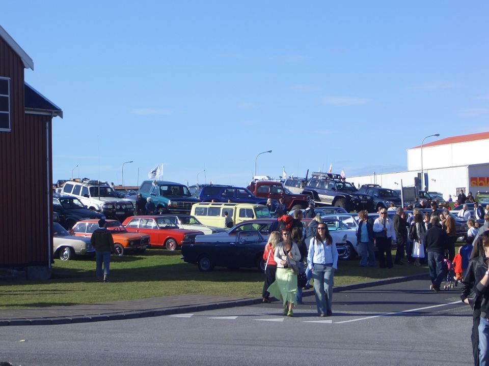 Outdoor classic car meet with vintage cars, lifted 4x4s, and crowds under a blue sky
