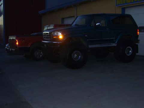 1996 Ford Bronco - A modified dark green Ford Bronco is parked next to a red pickup truck in front of a building.