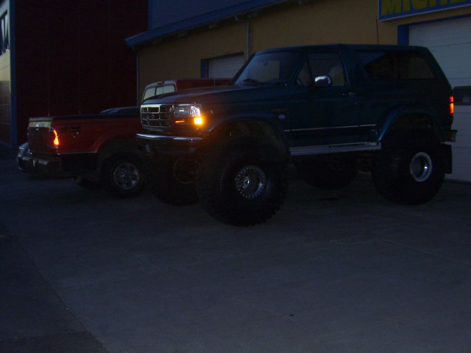 Lifted teal Ford Bronco on big tires beside a red pickup at dusk