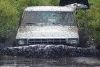 1990 Ford Bronco - This off-road vehicle navigates through a muddy obstacle, kicking up water.