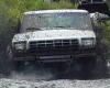 1990 Ford Bronco - A white Ford Bronco navigates a muddy terrain from a front-facing perspective.