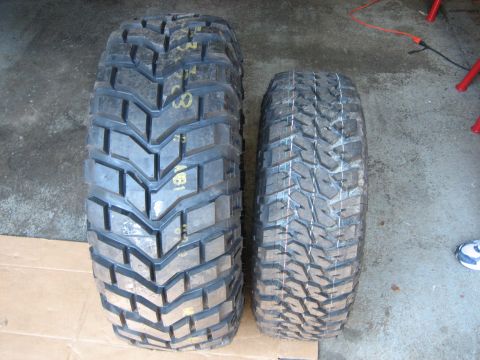 1990 Ford Bronco - Tread-face comparison of two off-road tires standing on a garage floor: a wide, deep-lugged mud tire with yellow paint markings on the left…