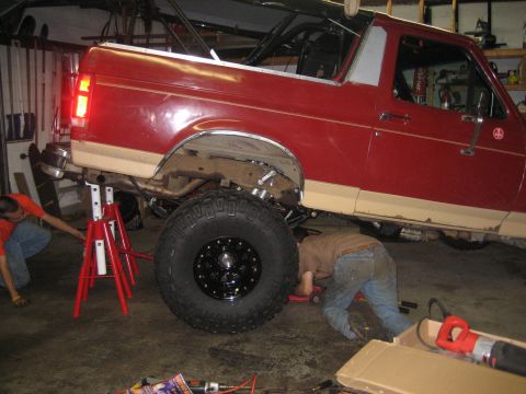 1990 Ford Bronco - Passenger-side view of a red-over-tan two-tone full-size Ford Bronco raised on red jack stands in a home garage, with the rear wheel remove…