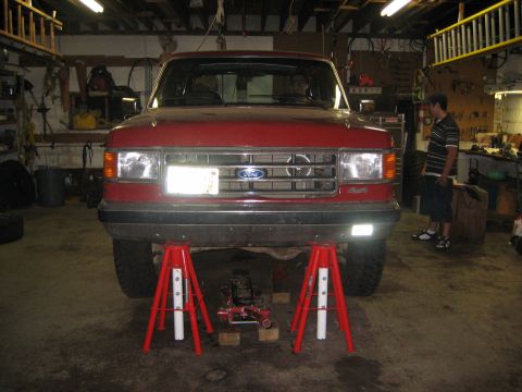 1990 Ford Bronco - Head-on front view of a red Ford Bronco raised on two tall red jack stands in a workshop, with a floor jack on wood blocks under the front…
