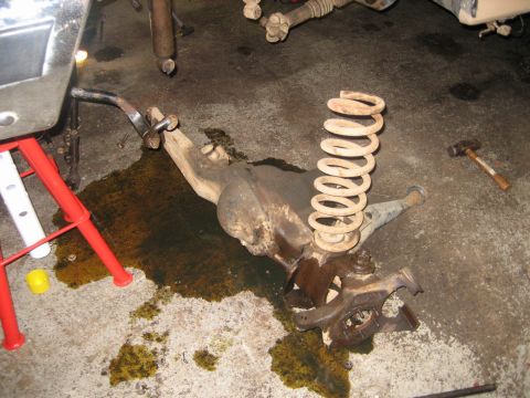 1990 Ford Bronco - A removed Dana-style front axle housing lying on a garage floor next to a spilled puddle of gear oil, still carrying its white-painted coil…