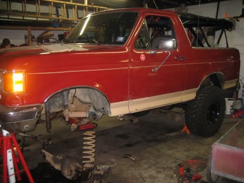 1990 Ford Bronco - Driver-side view of a red full-size Ford Bronco with the top removed, sitting on jack stands in a garage with the front wheel off and front…