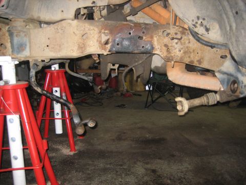1990 Ford Bronco - Under-truck view of a surface-rusted boxed frame rail and crossmember with the front axle out, taken from floor level between four red jack…