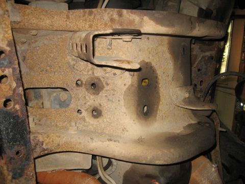 1990 Ford Bronco - Underside view of a heavily surface-rusted stamped-steel frame crossmember and floor/body mount area, shot from below in a dim garage. A sm…