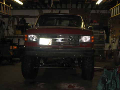 1990 Ford Bronco - Head-on view of a red lifted Ford Bronco inside a dim workshop with its headlights on, showing the Ford blue oval grille, 4x4 fender badge…