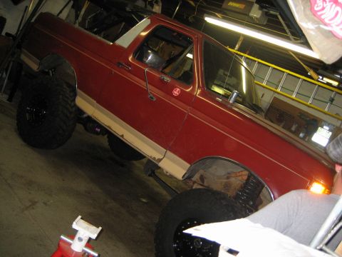 1990 Ford Bronco - Passenger side of a red and tan lifted Ford Bronco raised on a shop lift or jack stands inside a garage, top removed and doors on. The fron…