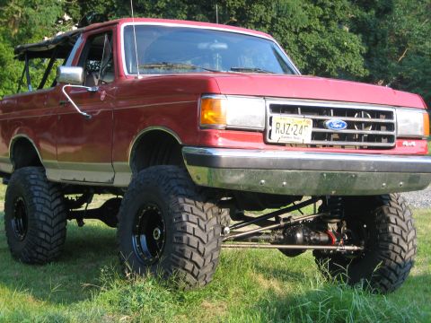 1990 Ford Bronco - A red and tan two-tone Ford Bronco lifted high on black steel wheels and oversized mud tires, parked on grass with its top removed and roll…