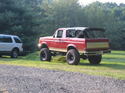 1990 Ford Bronco - Red and tan lifted Ford Bronco parked on grass beside a gravel drive, shot from the rear three-quarter with the hardtop removed and the sof…