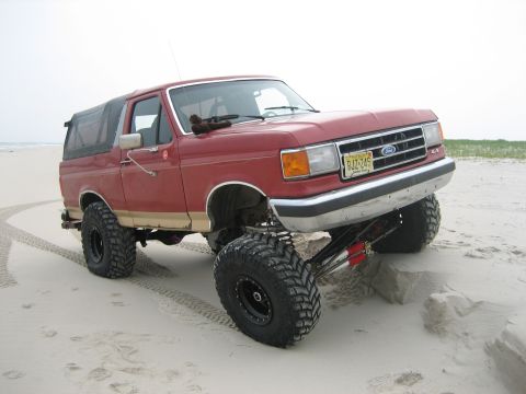 1990 Ford Bronco - Red and tan lifted Ford Bronco parked on a wide sand beach with tire tracks in the foreground and the ocean at left, viewed from the front…