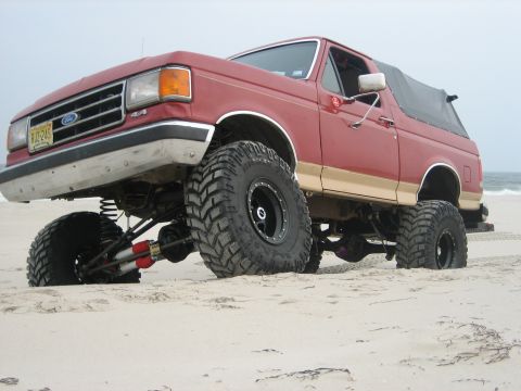 1990 Ford Bronco - Low front three-quarter view of a red and tan lifted Ford Bronco on a beach with the ocean behind it, articulating over the sand so the dri…