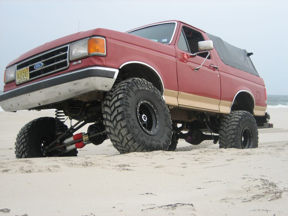 Red lifted Ford Bronco on beach sand with front tire lifted, ocean in background