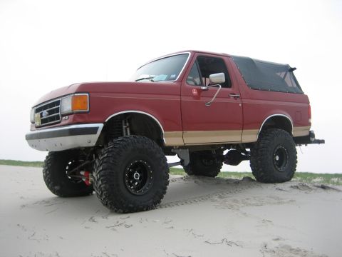 1990 Ford Bronco - Driver-side three-quarter front view of a lifted red-and-tan full-size Ford Bronco parked on a sand dune under an overcast sky, wearing a b…