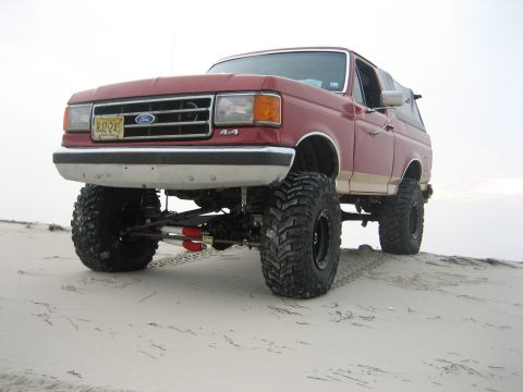 1990 Ford Bronco - Low front three-quarter shot of a lifted red-and-tan full-size Ford Bronco standing on a sand dune, showing the exposed solid front axle, r…