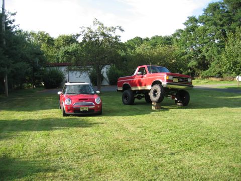1990 Ford Bronco - Size-contrast shot on a sunny lawn: a lifted red full-size Ford Bronco on tall mud-terrain tires and black wheels parked beside a red-with-…