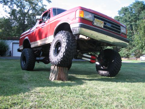 1990 Ford Bronco - Low front three-quarter shot of a lifted red and silver two-tone Ford SUV flexing on a grass lawn with its left front tire perched on a tre…