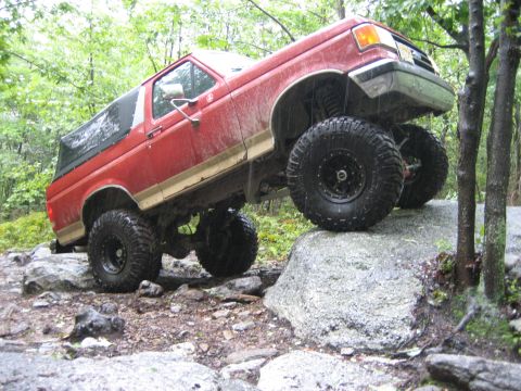 1990 Ford Bronco - Red and tan two-tone lifted Ford Bronco climbing a wet granite ledge on a wooded trail, passenger side flexed with the front tire lifted of…