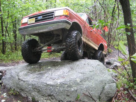 1990 Ford Bronco - Low front three-quarter shot of a red lifted Ford Bronco flexed over a large granite boulder on a wet wooded trail, with the driver-side fr…