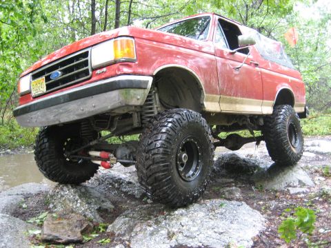 1990 Ford Bronco - This red 1990 Ford Bronco is equipped with a lifted suspension and large off-road tires, navigating a rocky terrain.