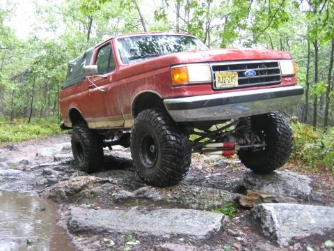 1990 Ford Bronco - Red 1990 Ford Bronco tackling an off-road trail with large tires.