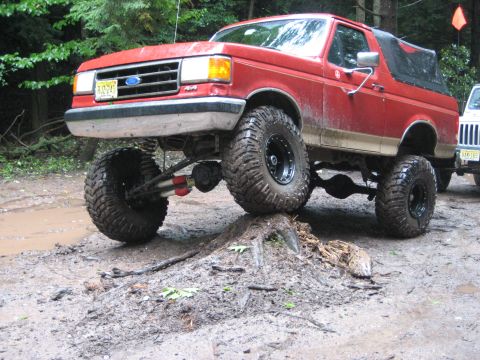 1990 Ford Bronco - A modified red Ford Bronco navigates an obstacle on a muddy trail, with another vehicle in the background.