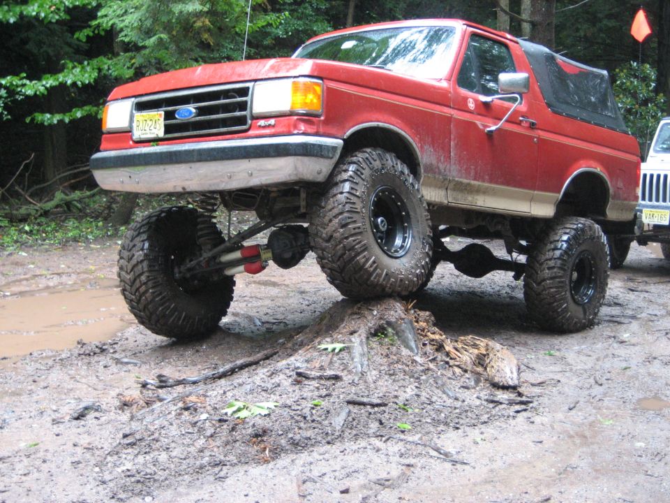 Red lifted Ford Bronco on a dirt mound, front right tire elevated. Muddy terrain, trees in background. Another vehicle visible to the right