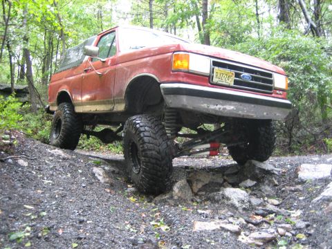 1990 Ford Bronco - Red lifted Ford Bronco crawling down a rocky, rain-soaked trail through green woods, shot from the front three-quarter with the front axle…