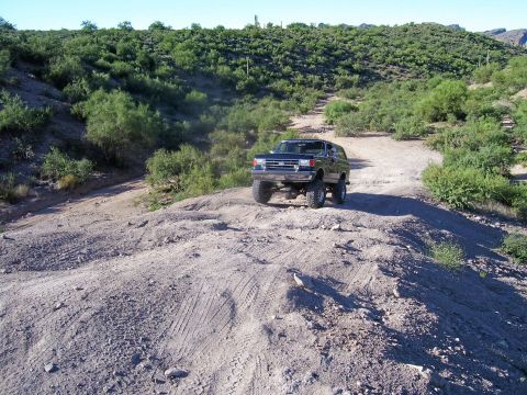 1991 Ford Bronco - A lifted black Ford Bronco navigates a dusty, arid terrain with sparse vegetation under a clear blue sky.