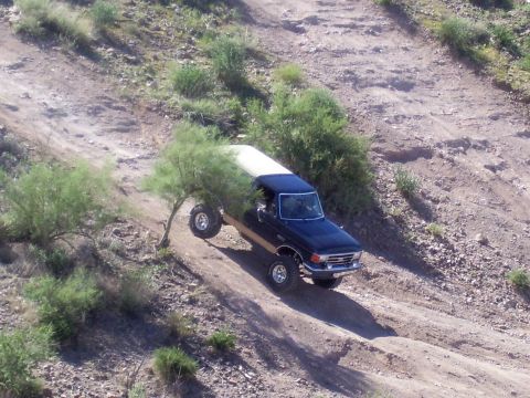 1991 Ford Bronco - A dark blue 1987 Ford Bronco with a white top navigates a steep, dusty off-road trail with its front passenger wheel elevated.