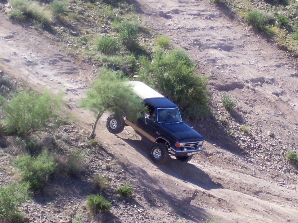 Dark blue 1987 Ford Bronco with a white top driving off-road on a dirt hill, front passenger side wheel lifted