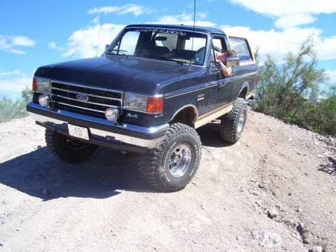 1991 Ford Bronco - This lifted Ford Bronco is navigating a dusty, off-road trail under a bright blue sky.