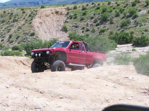 1991 Ford Bronco - A red pickup truck with large tires and a roll bar drives off-road in a desert landscape.