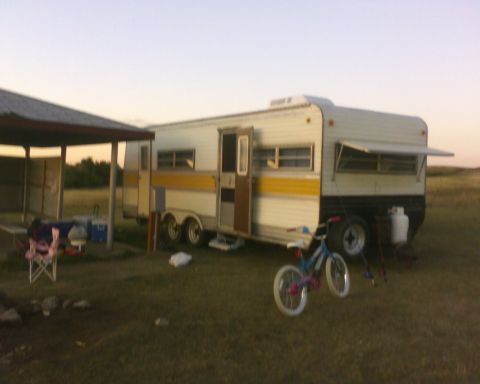 1989 Ford Bronco - A camper trailer is parked in a grassy area next to a covered shelter with outdoor furniture.